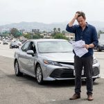 A middle-aged man holding paperwork stands stressed next to his damaged car on a California highway after an accident, illustrating a blog post about medical bills exceeding policy limits. no text, no watermark