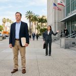 Man holding legal documents standing outside a California courthouse while a lawyer gestures toward the entrance, representing partial fault car accident compensation claims in California.